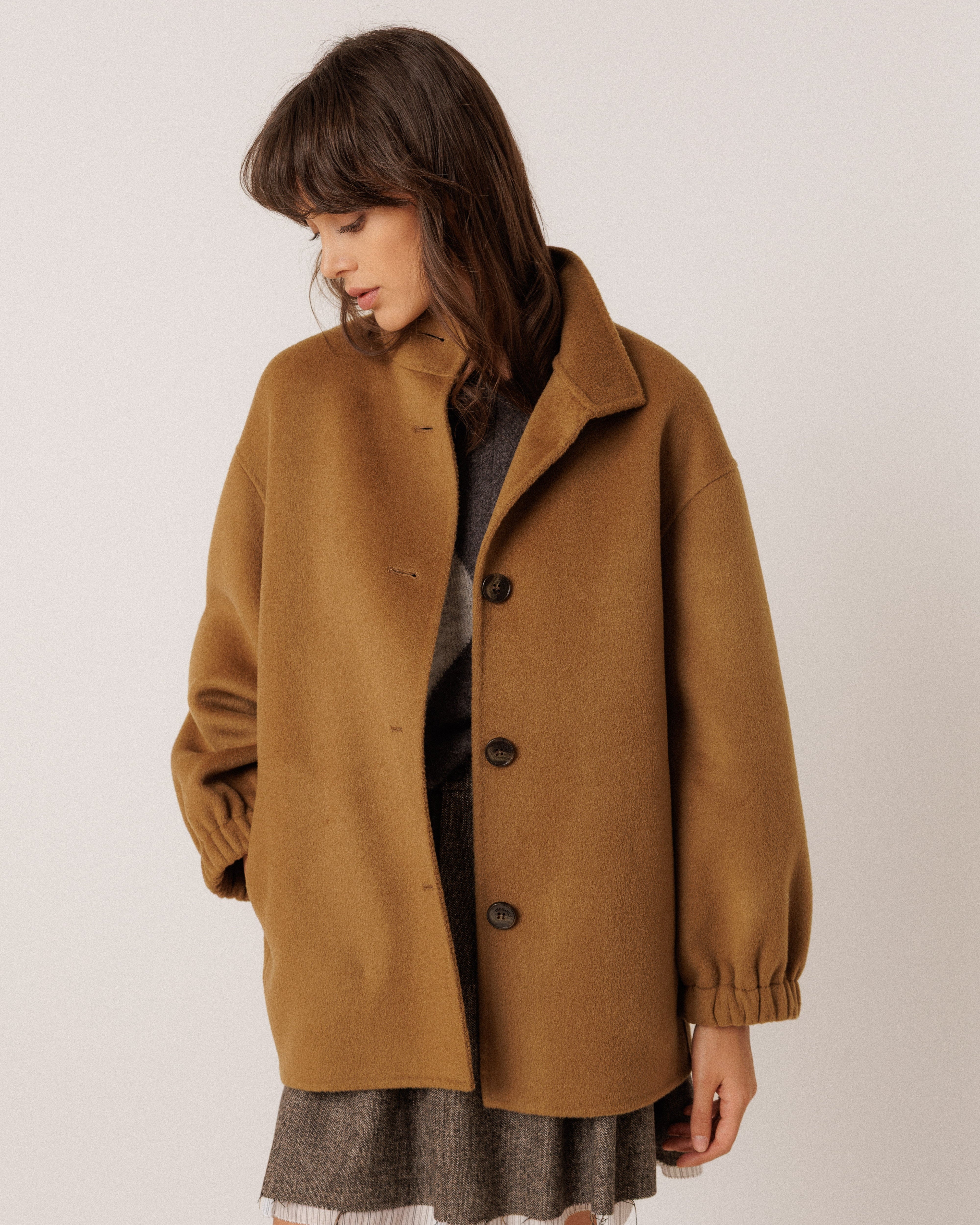 A woman wearing an oversized camel brown wool-blend coat with dark buttons, styled over a dark top and a gray pleated skirt, photographed against a plain light gray studio background.