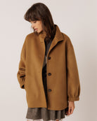 A woman wearing an oversized camel brown wool-blend coat with dark buttons, styled over a dark top and a gray pleated skirt, photographed against a plain light gray studio background.