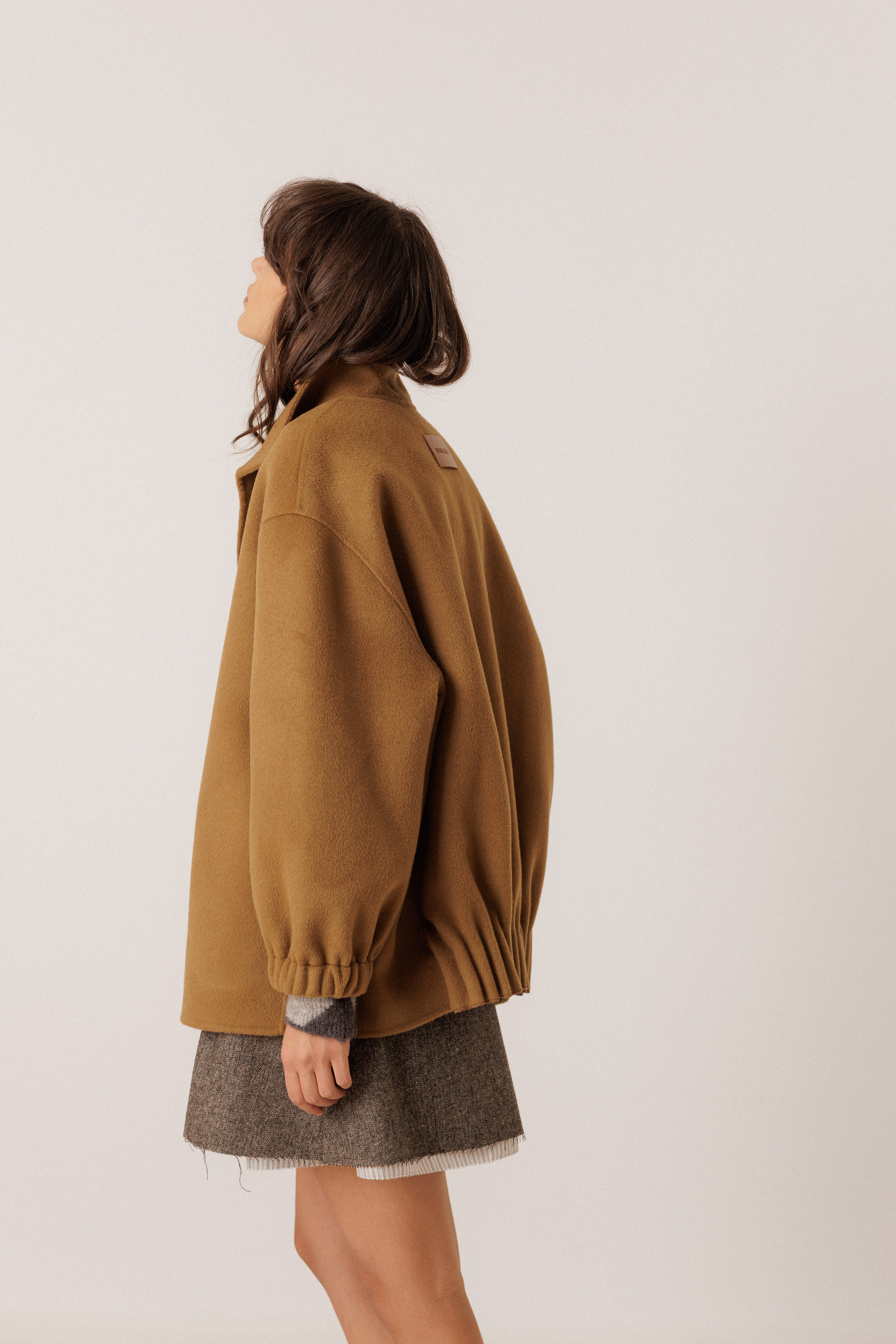 A woman shown from the side profile wearing an oversized camel brown jacket and a gray textured skirt, photographed against a plain light gray studio background.