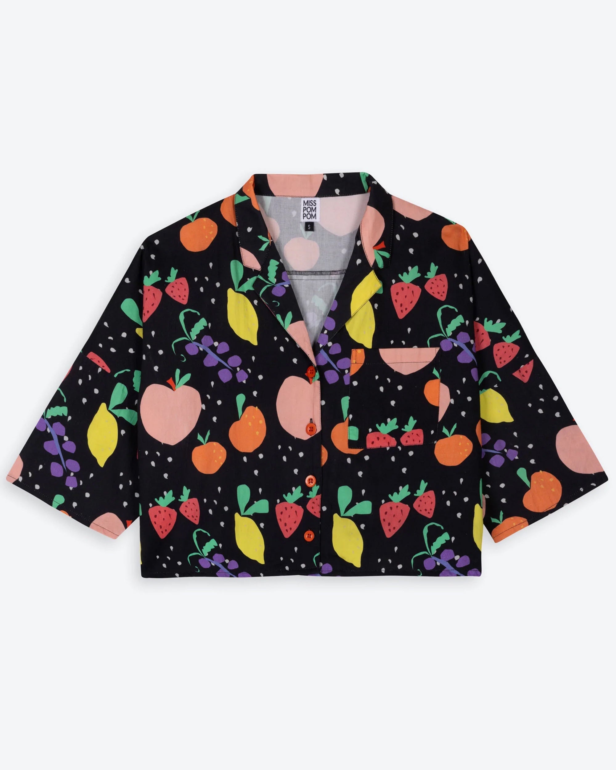 A cropped, boxy black shirt with a playful fruit print, including peaches, lemons, strawberries, grapes, and oranges, set against a black background with white dots. The shirt has a collar, red buttons, and a chest pocket with a fruit print.