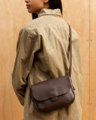 A person wearing a tan jacket and patterned pants carries a dark brown pebbled leather crossbody bag with a flap closure and thin strap, standing against a light wood plank wall.
