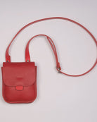 A small red pebbled leather crossbody bag with a long matching shoulder strap, laid flat on a light off-white background.