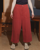 A model wearing a dark navy t-shirt and high-waisted, wide-leg red corduroy pants, standing in a modern indoor setting with wooden furniture and a hanging lamp.