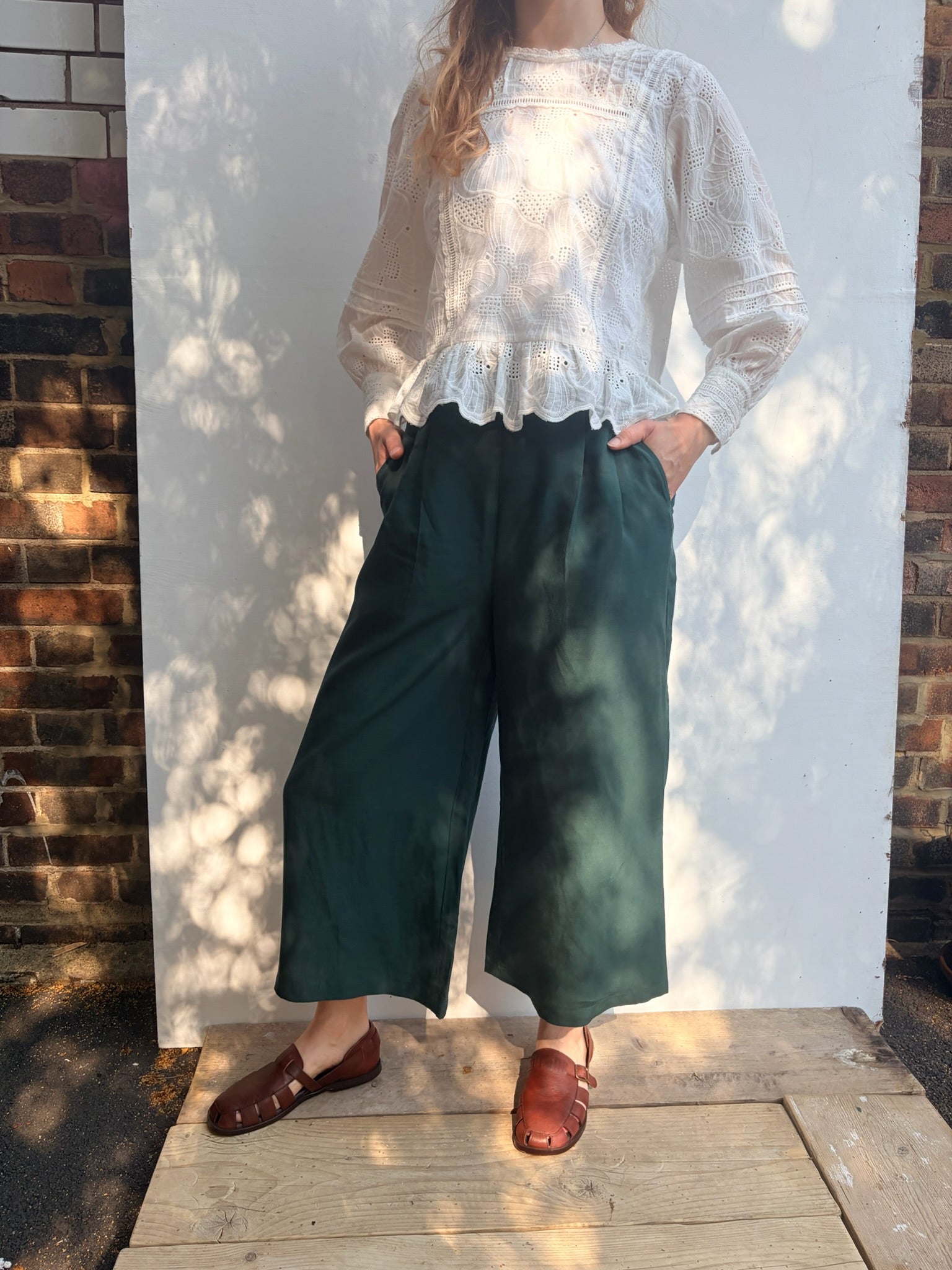 A person wearing a white long-sleeve lace-trim blouse and dark green wide-leg cropped pants, standing outdoors against a white wall with brick sides and dappled sunlight.