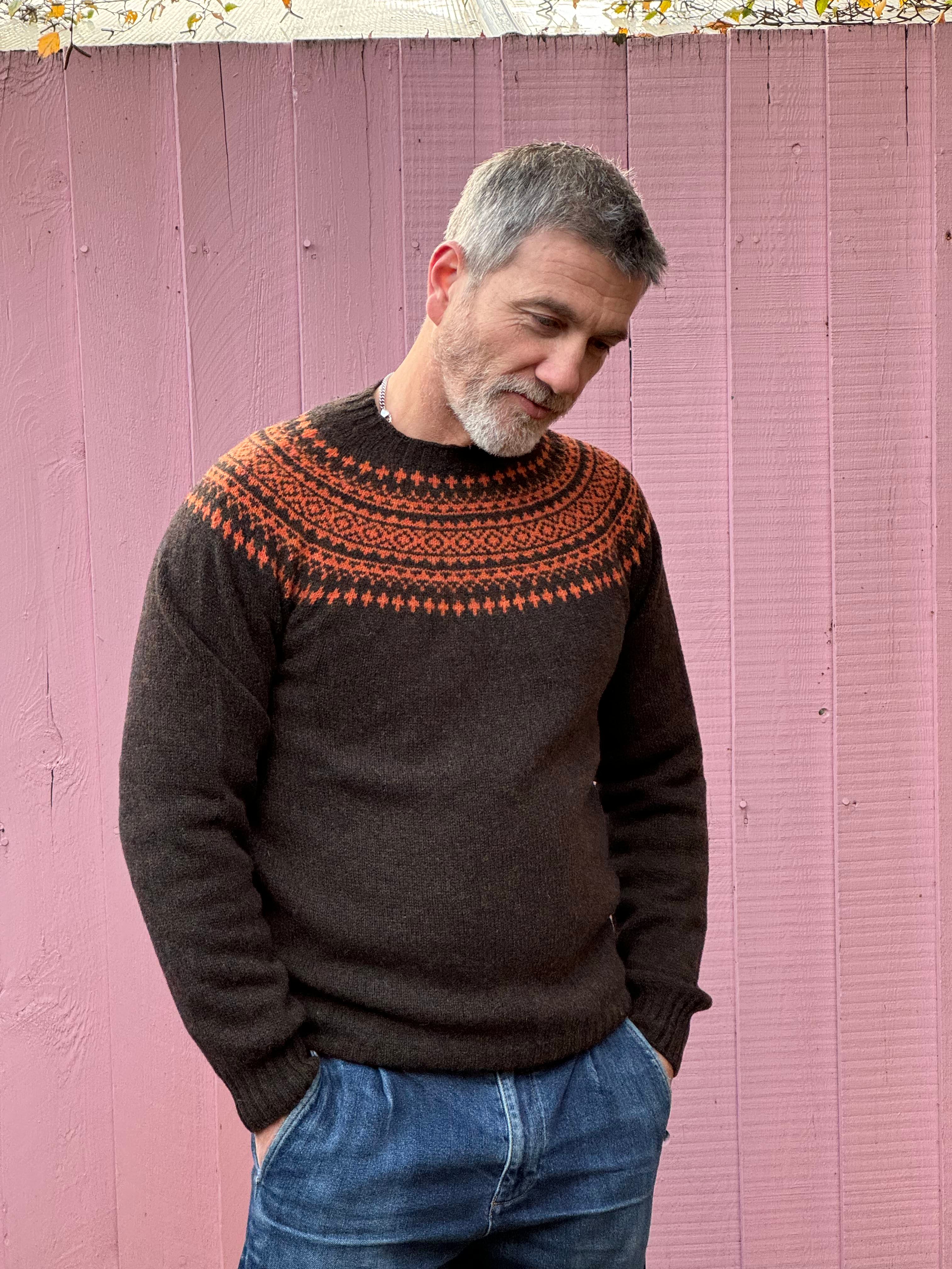A man wearing a chocolate brown and orange fairisle pattern jumper with a ribbed round neck and long sleeves.