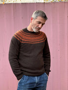 A man wearing a chocolate brown and orange fairisle pattern jumper with a ribbed round neck and long sleeves.