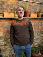 A man wearing a chocolate brown and orange fairisle pattern jumper with a ribbed round neck and long sleeves.