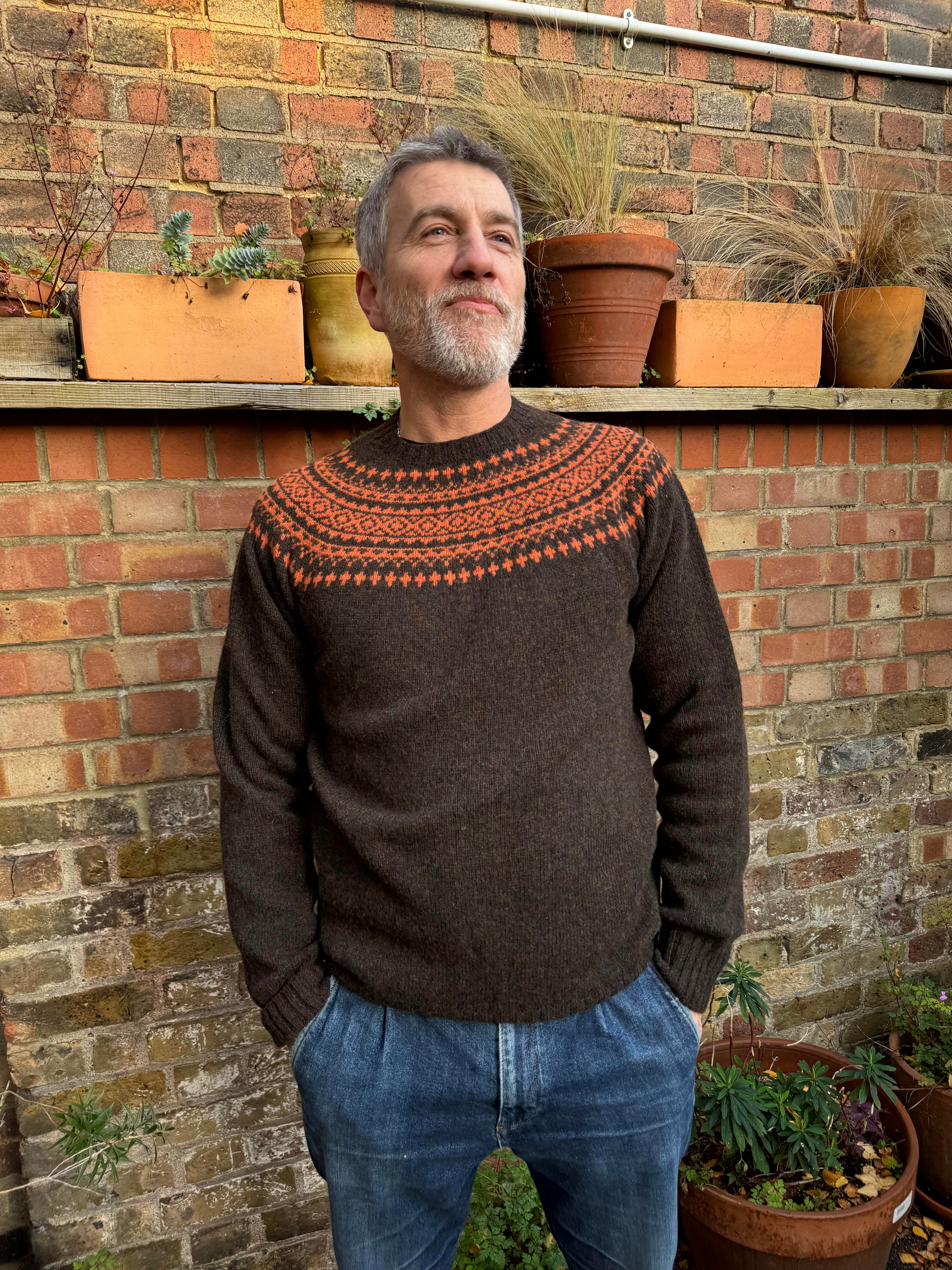A man wearing a chocolate brown and orange fairisle pattern jumper with a ribbed round neck and long sleeves.