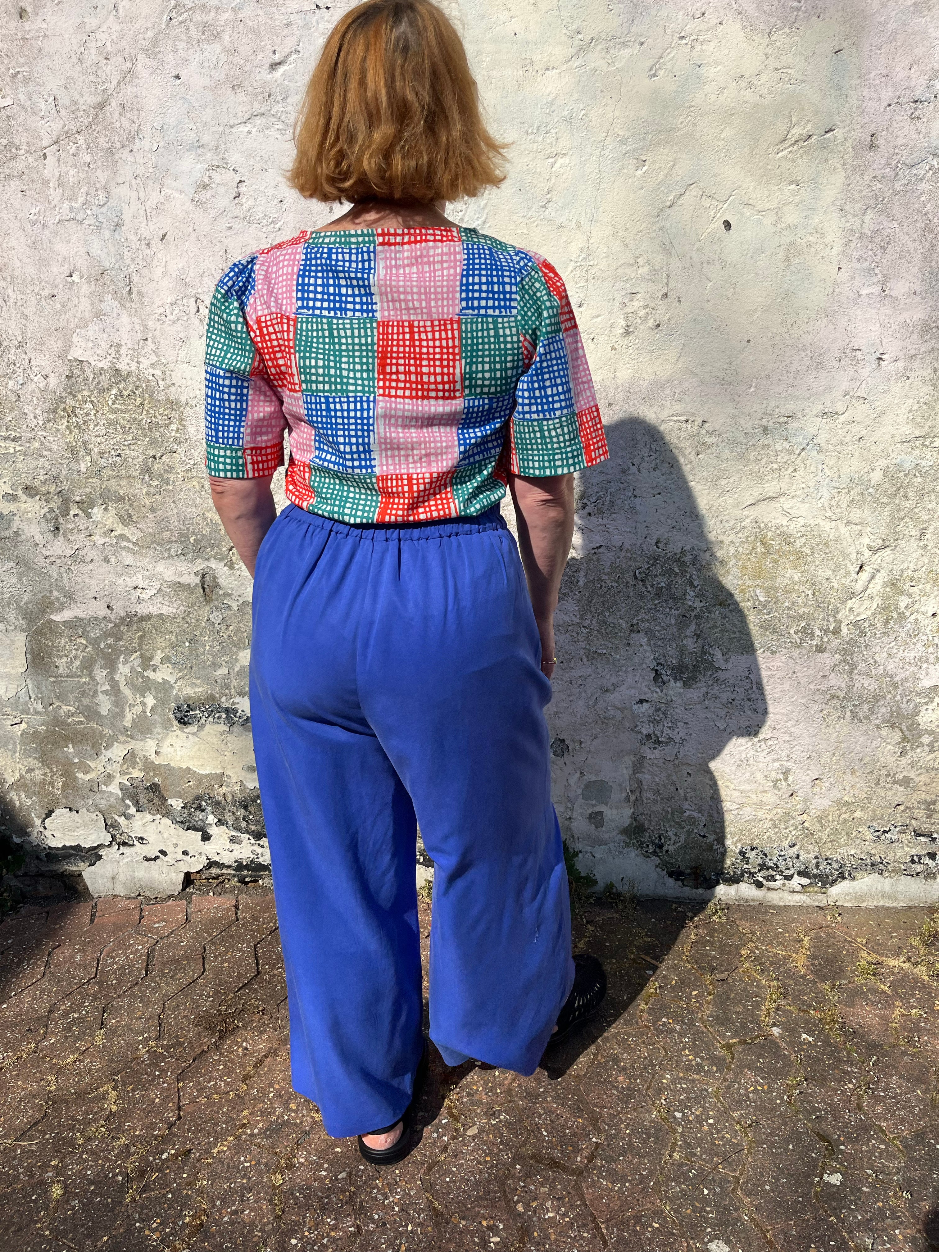A person standing against a wall wearing cobalt lyocell wide-leg trousers with a flat front waistband, elasticated back, and pleats at the front.