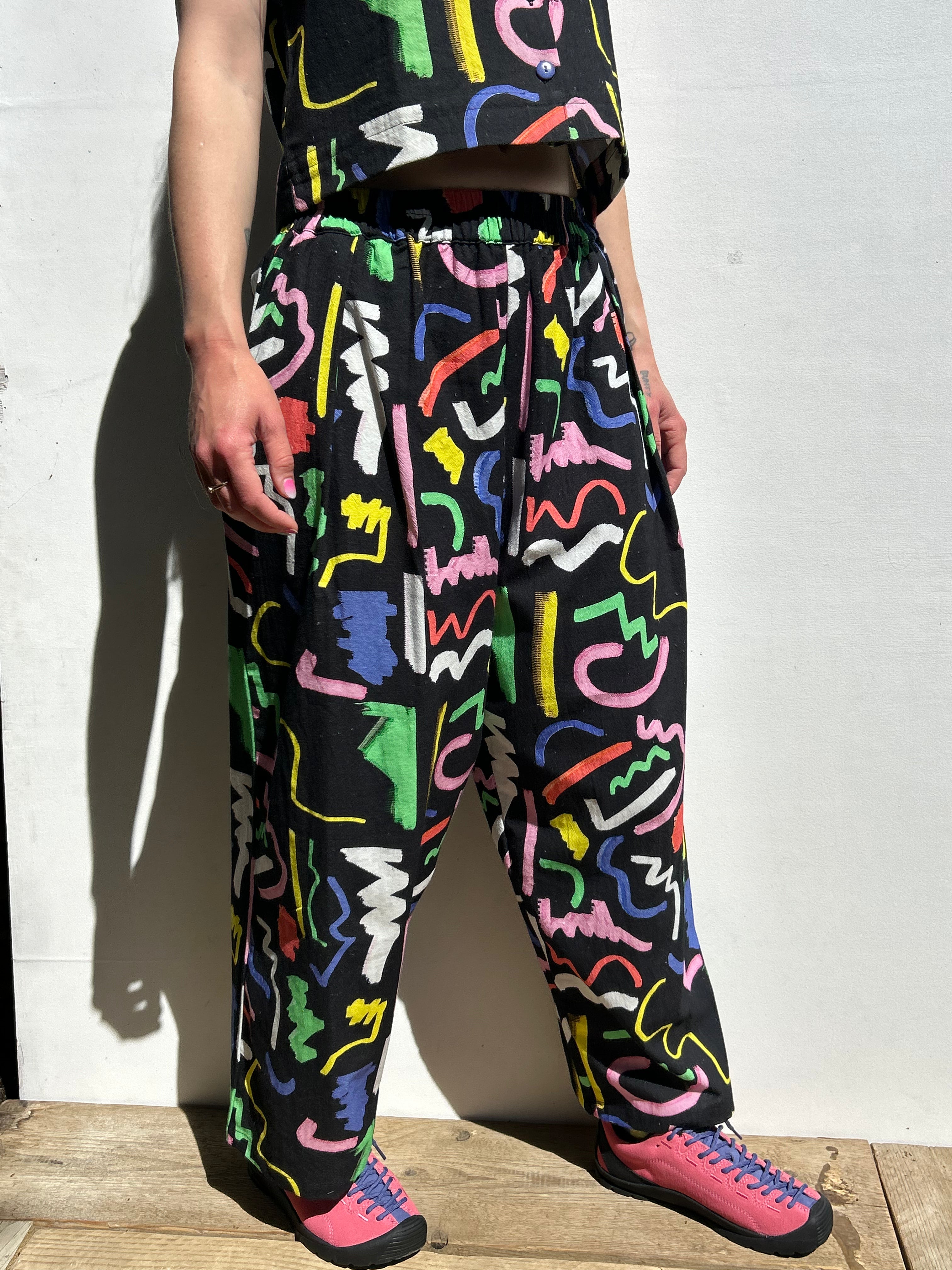 Person wearing colorful abstract pattern pants and pink shoes against a white wall.