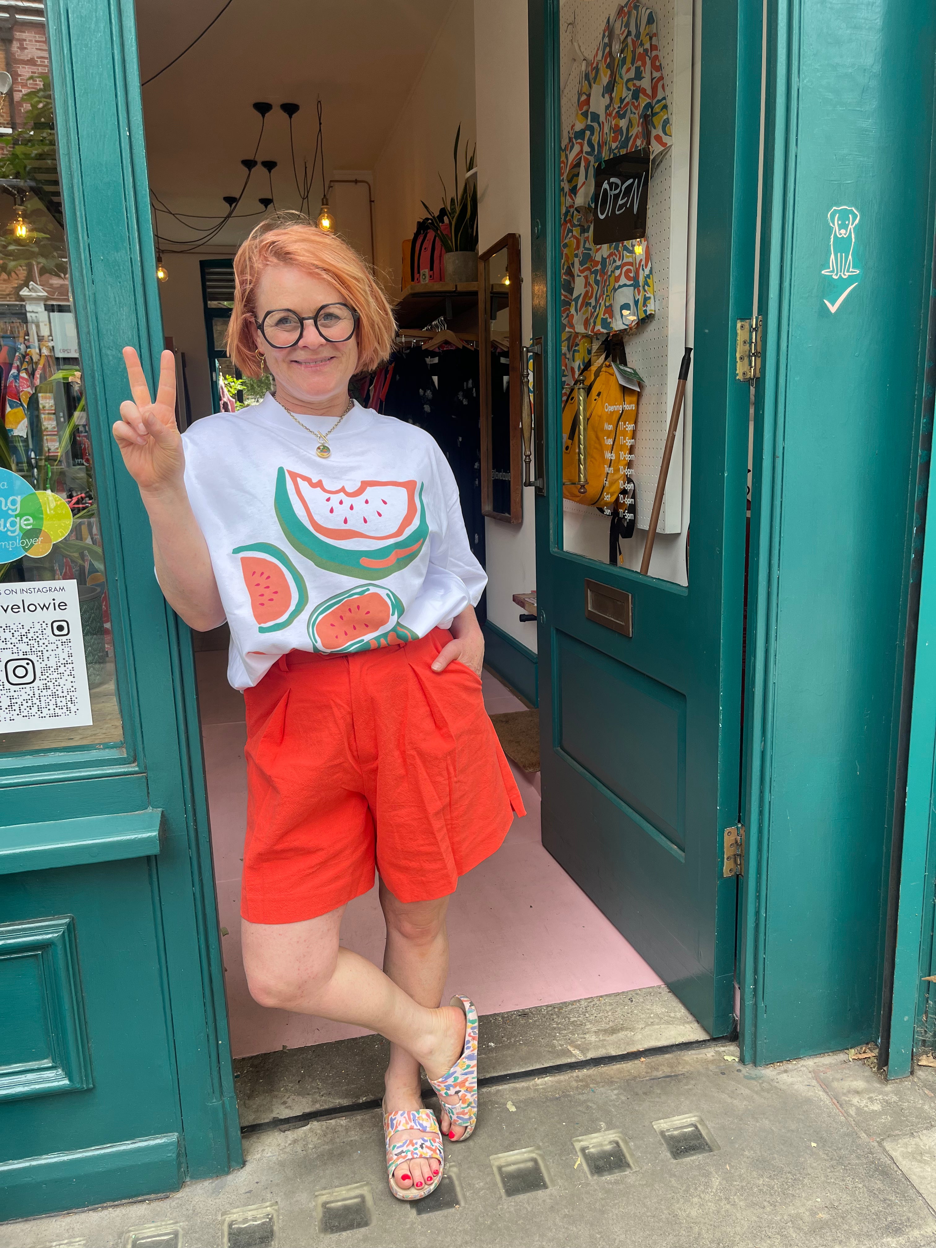Person wearing a white t-shirt with a watermelon design and red shorts standing in front of a teal door.