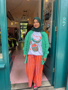 Woman standing in a doorway wearing a green cardigan, white shirt with graphic design, and orange striped pants.