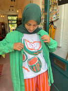 Person wearing a green hijab, white t-shirt with watermelon design, and orange pants in a store setting.