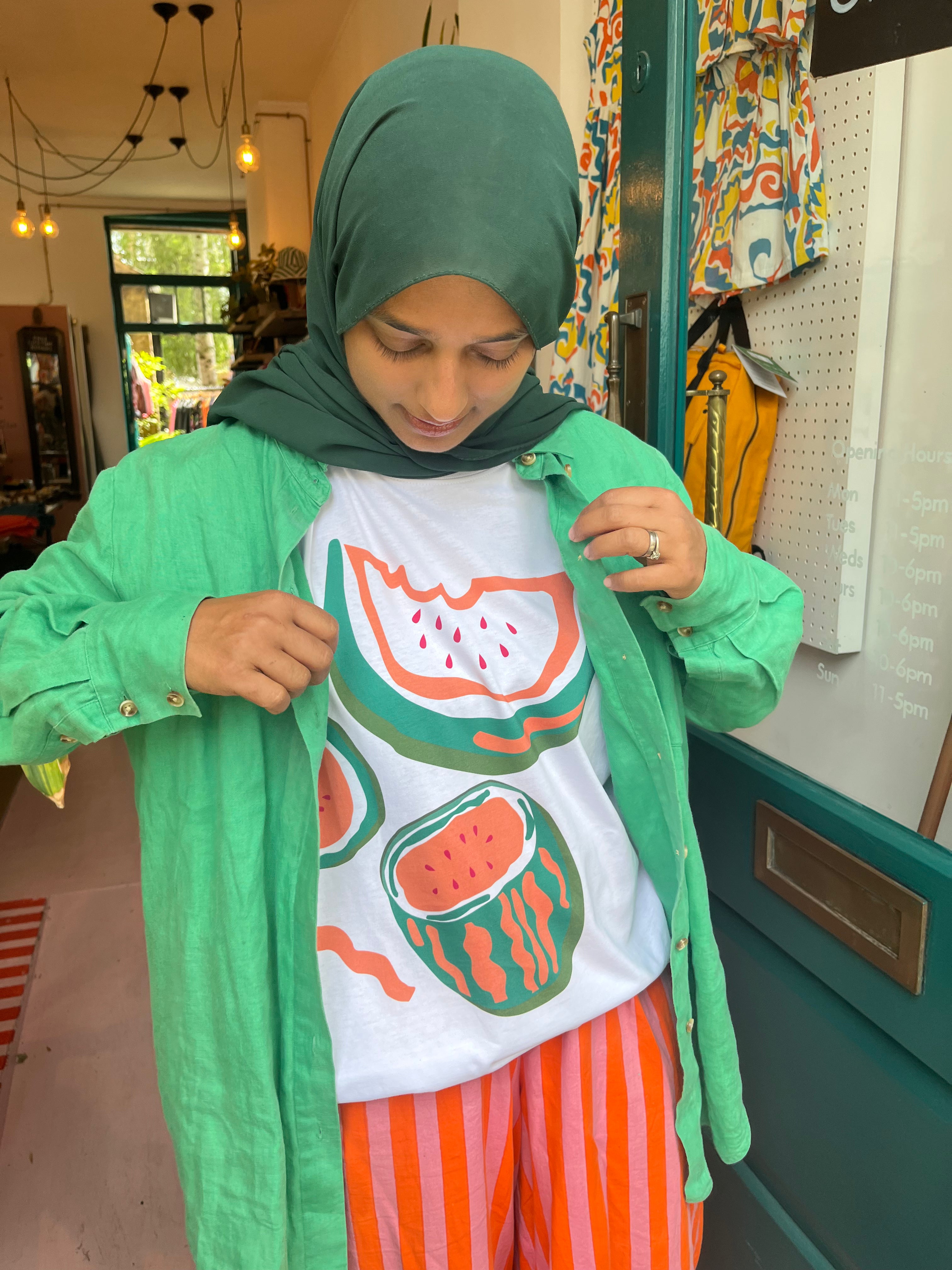 Person wearing a green hijab, white t-shirt with watermelon design, and orange pants in a store setting.