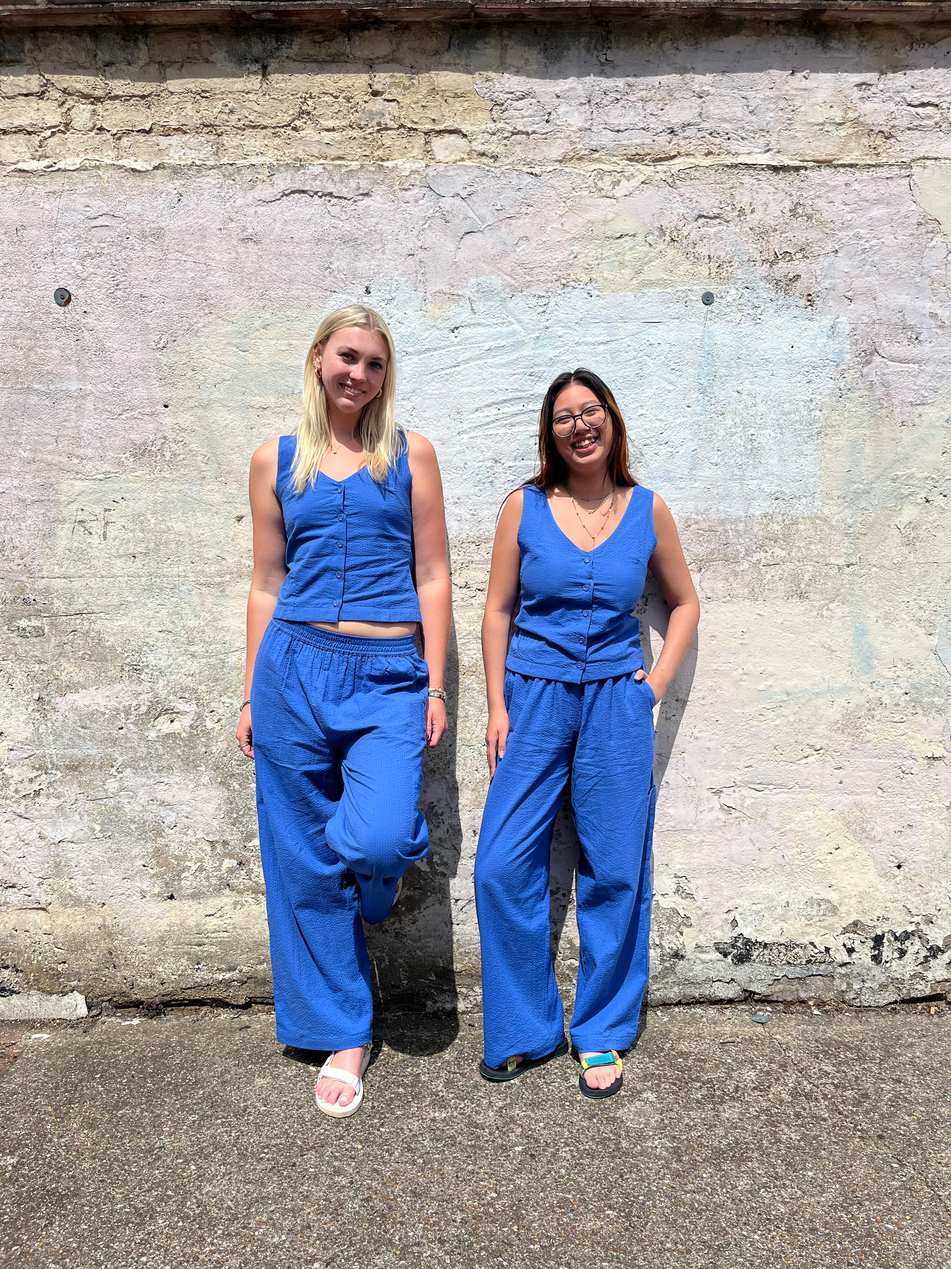 Two women wearing matching blue jumpsuits standing against a textured wall.