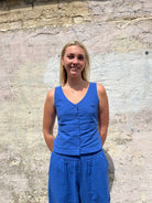 Woman wearing a blue sleeveless top and pants standing against a stone wall.