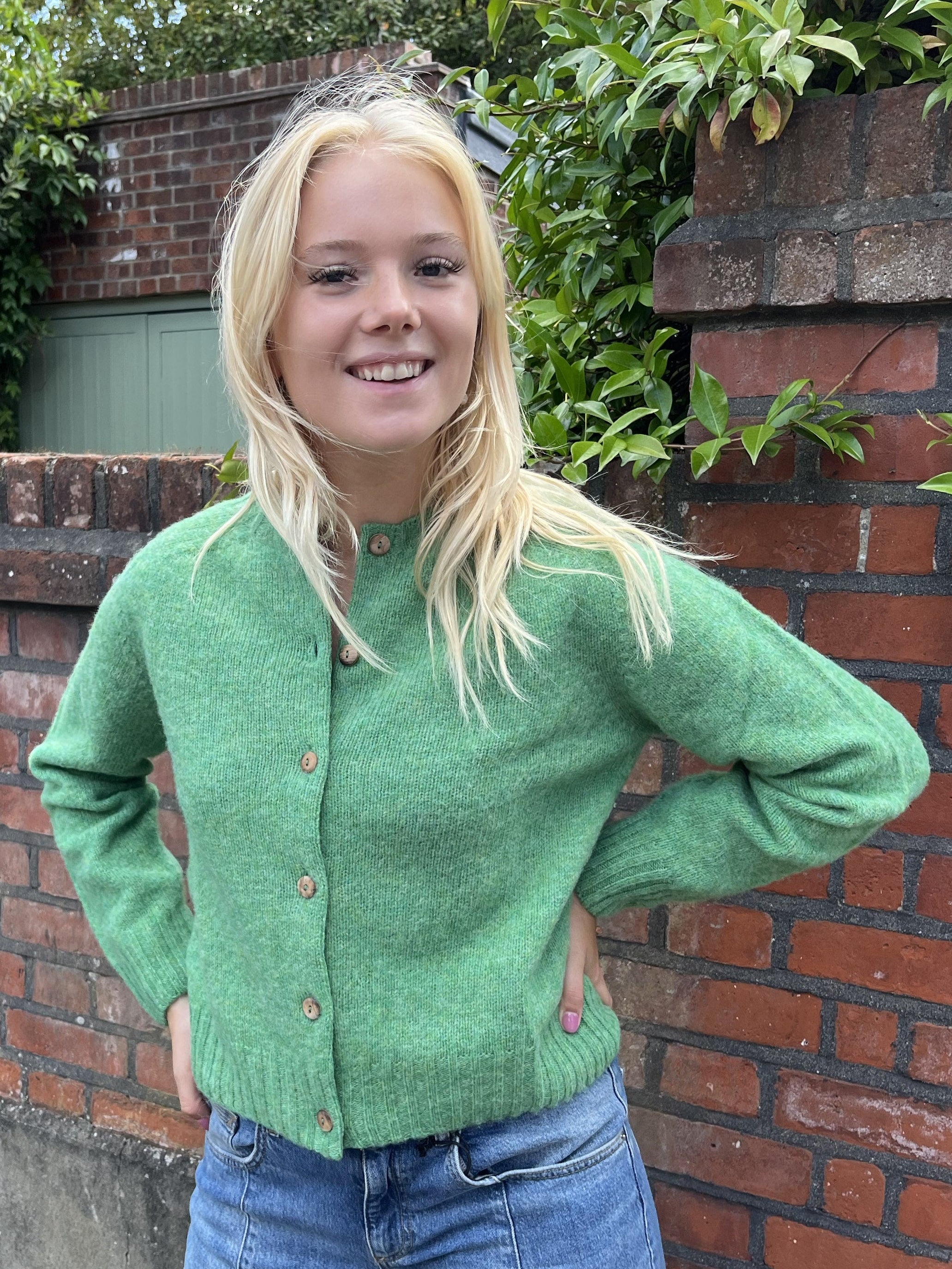 Person wearing a green cardigan standing in front of a brick wall with plants.
