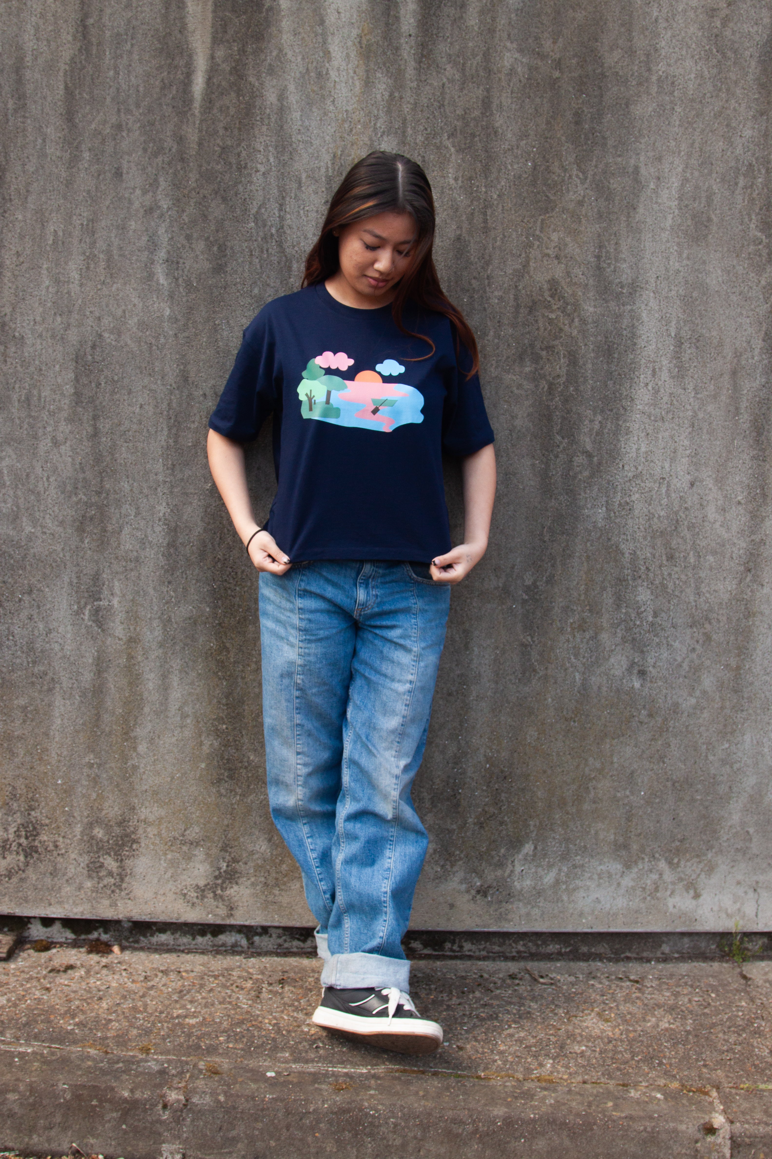 A navy blue t-shirt with a graphic print of a lake scene at dusk with a lone canoe, pink and blue waters, and green trees.