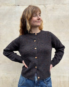 A woman wearing a dark brown textured knit cardigan with wooden buttons, paired with blue jeans, standing against a light beige stone wall.