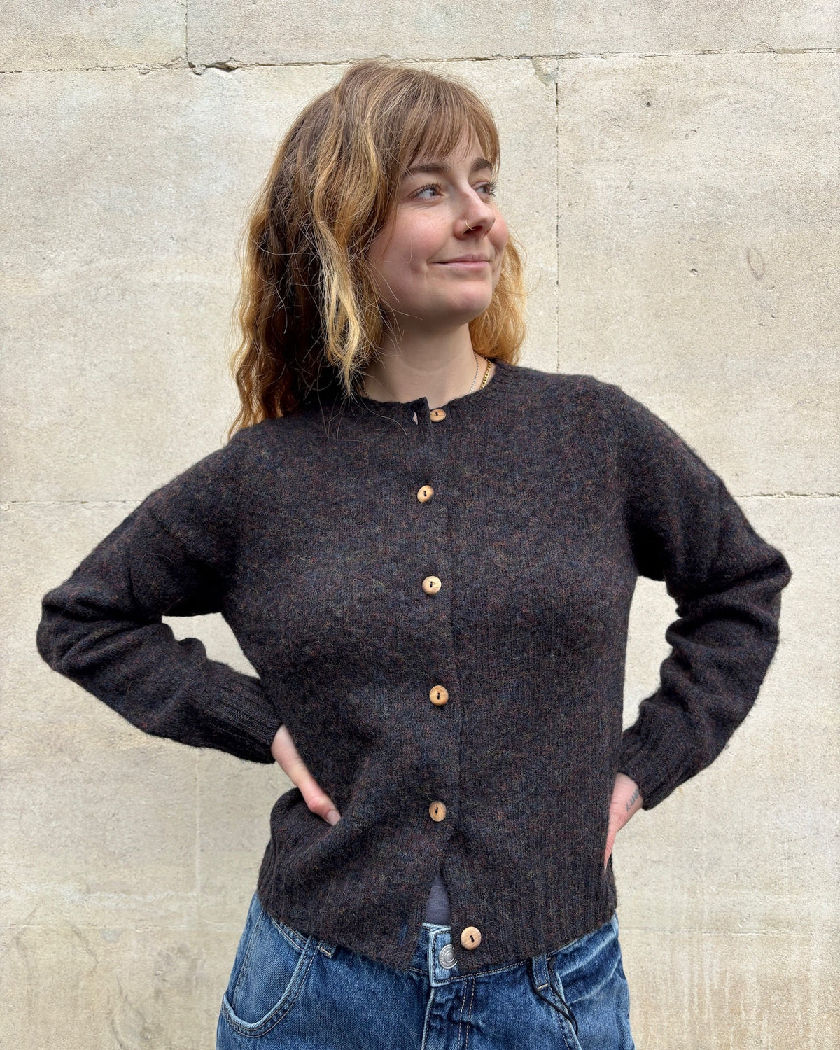 A woman wearing a dark brown textured knit cardigan with wooden buttons, paired with blue jeans, standing against a light beige stone wall.