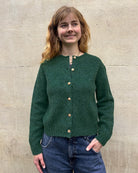A woman with shoulder-length light brown hair stands against a light beige stone wall, wearing a dark green knit button-front cardigan with wooden buttons and blue jeans.