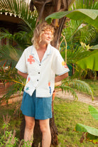 Person wearing a white shirt with colorful designs and blue shorts standing in a tropical setting.