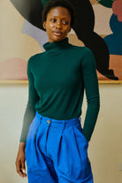 Person wearing a green turtleneck and blue pants against a colorful abstract wall.