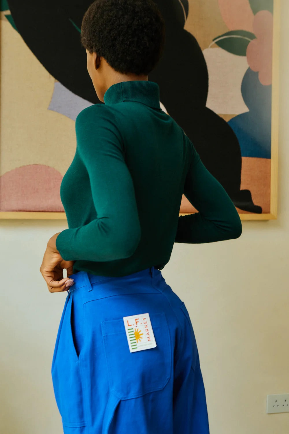 Person wearing a green sweater and blue pants with a colorful abstract painting in the background.