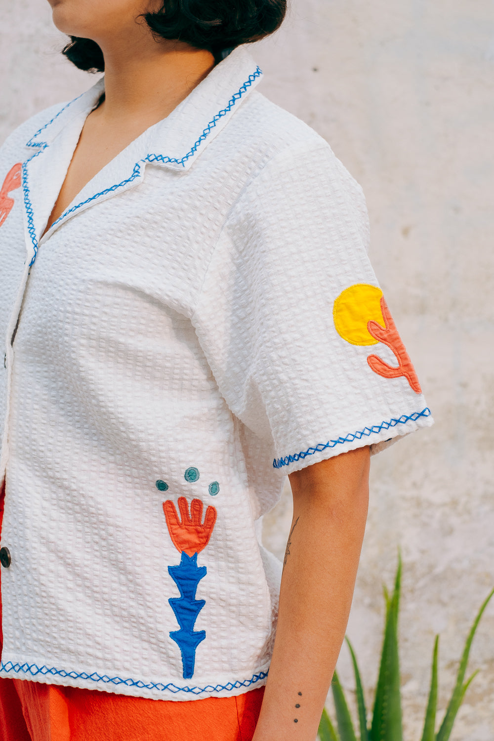 A white cotton blouse with colorful hand embroidery details, featuring a notched collar, wide cut sleeves, and a boxy fit.
