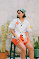 A white cotton blouse with colorful hand embroidery details, featuring a notched collar, wide cut sleeves, and a boxy fit.