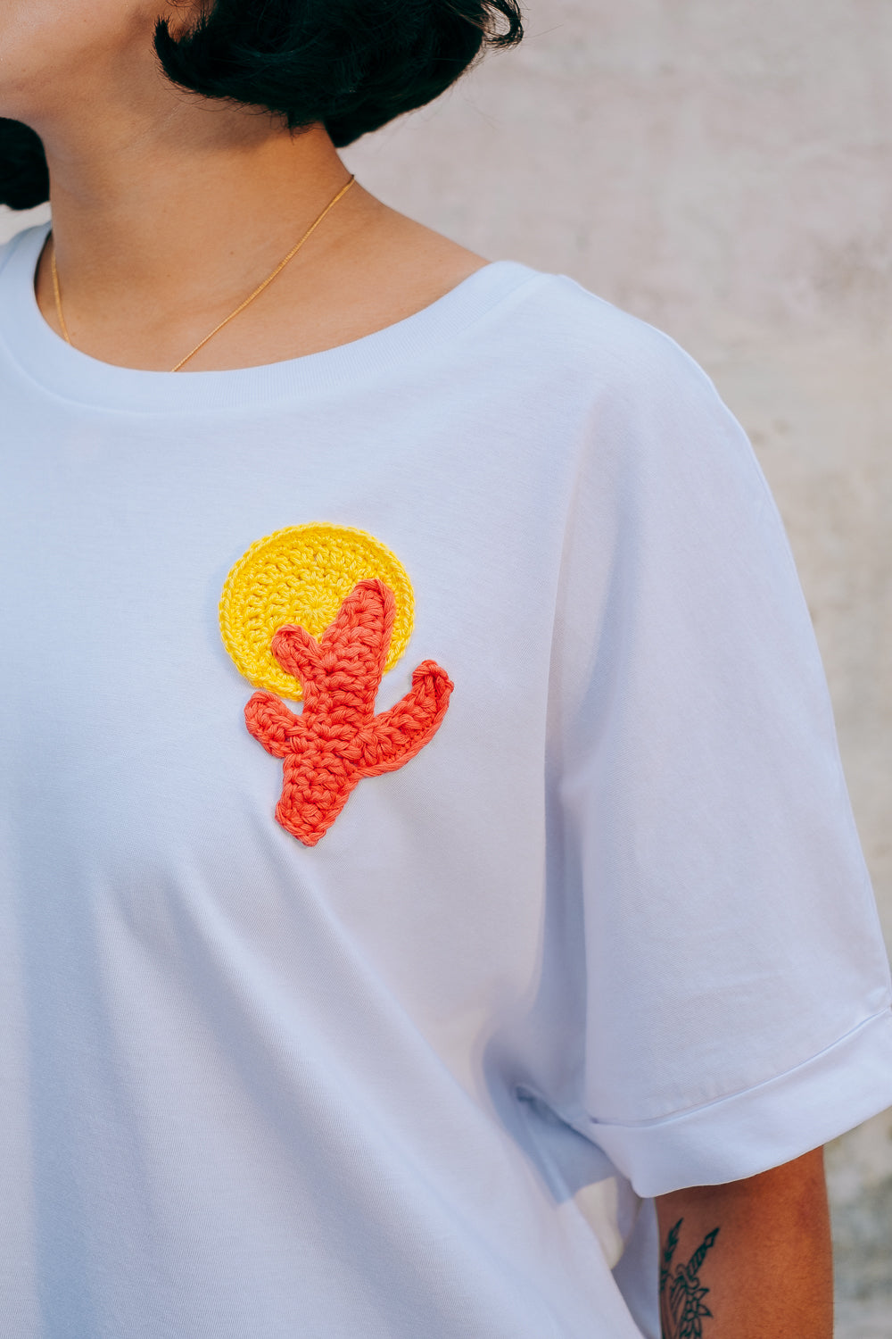A person wearing a white crew neck t-shirt with a hand-crocheted red and yellow cactus and sun motif, paired with a blue skirt and a red belt.