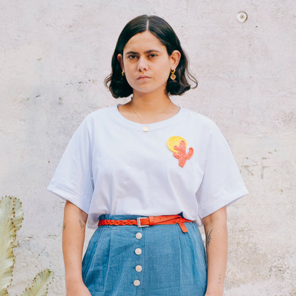A person wearing a white crew neck t-shirt with a hand-crocheted cactus and sun design, paired with a blue skirt and an orange belt.