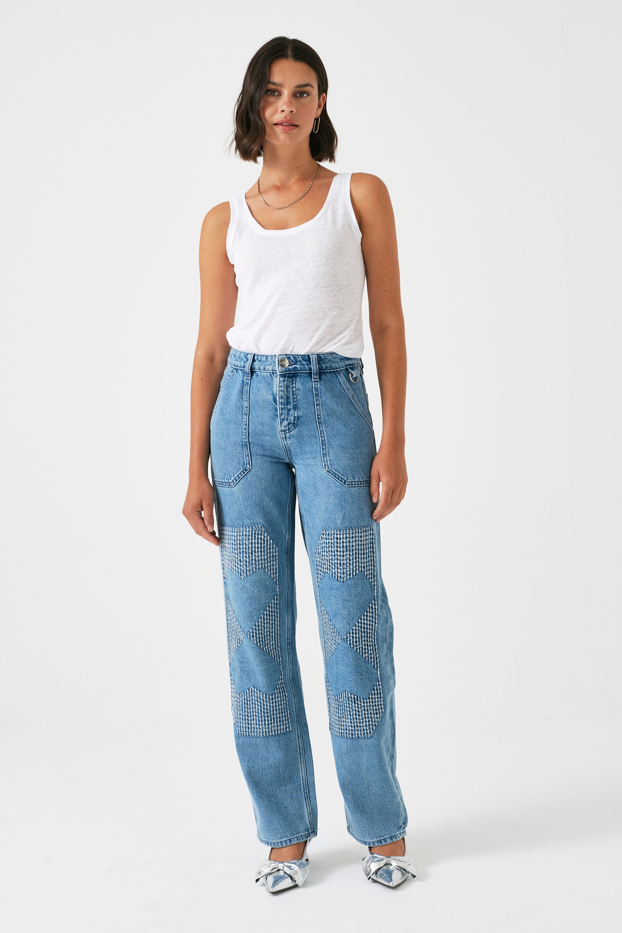 A pair of blue carpenter style jeans with embroidery on the front legs and a center front seam, displayed against a white background.