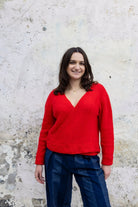 Woman wearing a red sweater and blue pants standing against a textured white wall.
