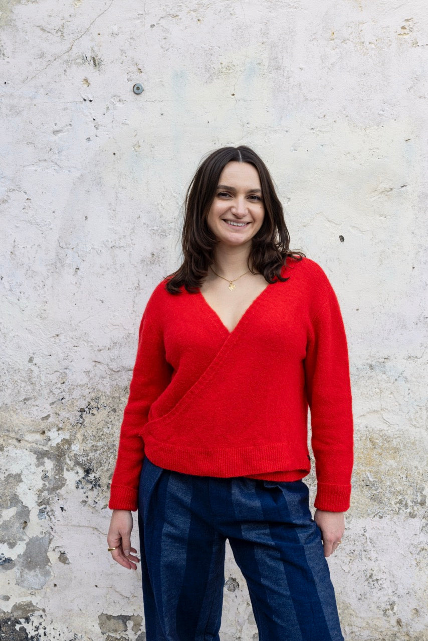 Woman wearing a red sweater and blue pants standing against a textured white wall.