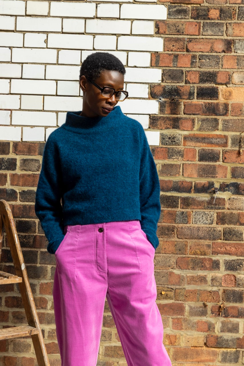 Person wearing a blue sweater and pink pants standing against a brick wall.