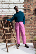 Person wearing a blue sweater and pink pants standing next to a wooden ladder against a brick wall.