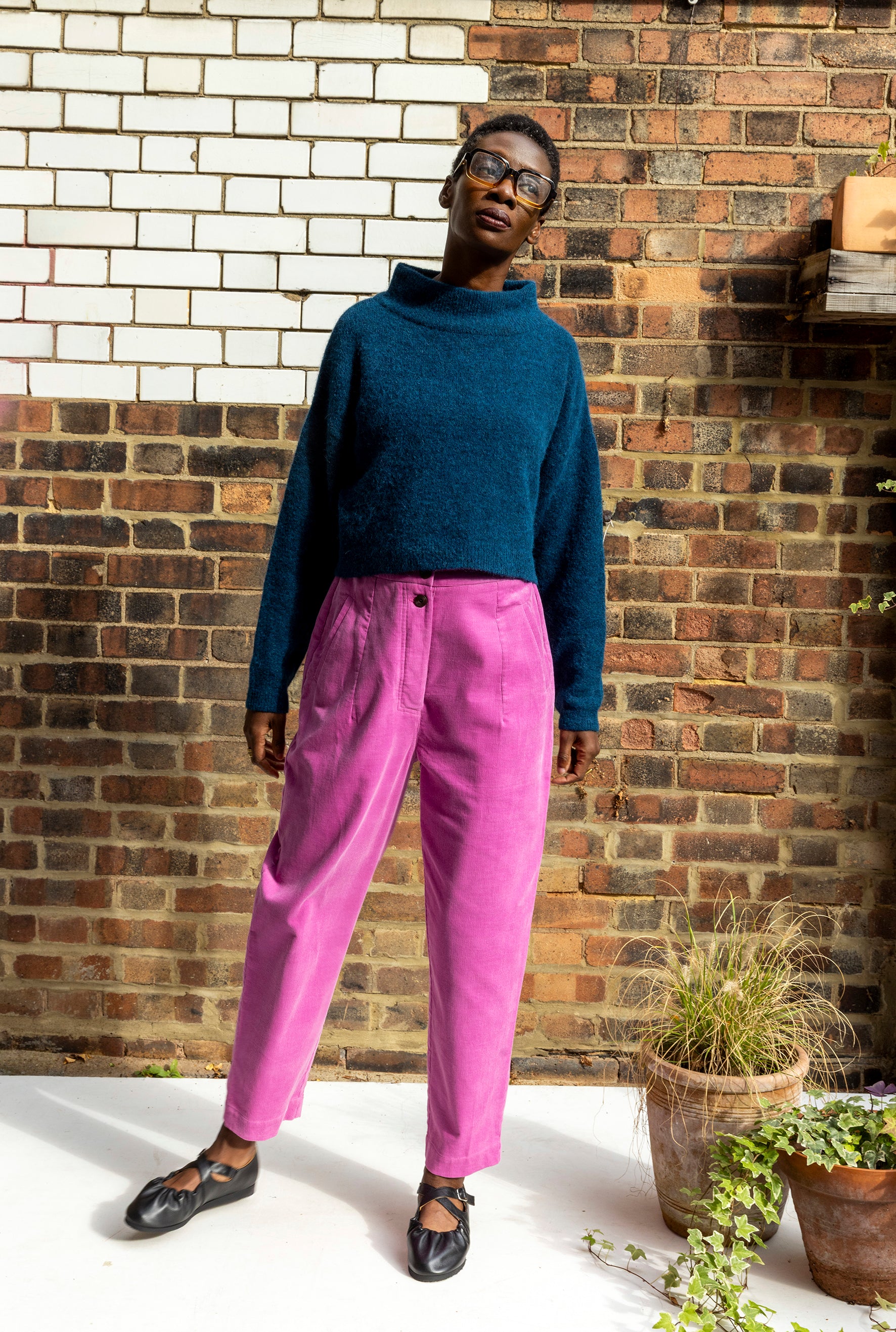 Person wearing a blue sweater and pink pants standing against a brick wall.