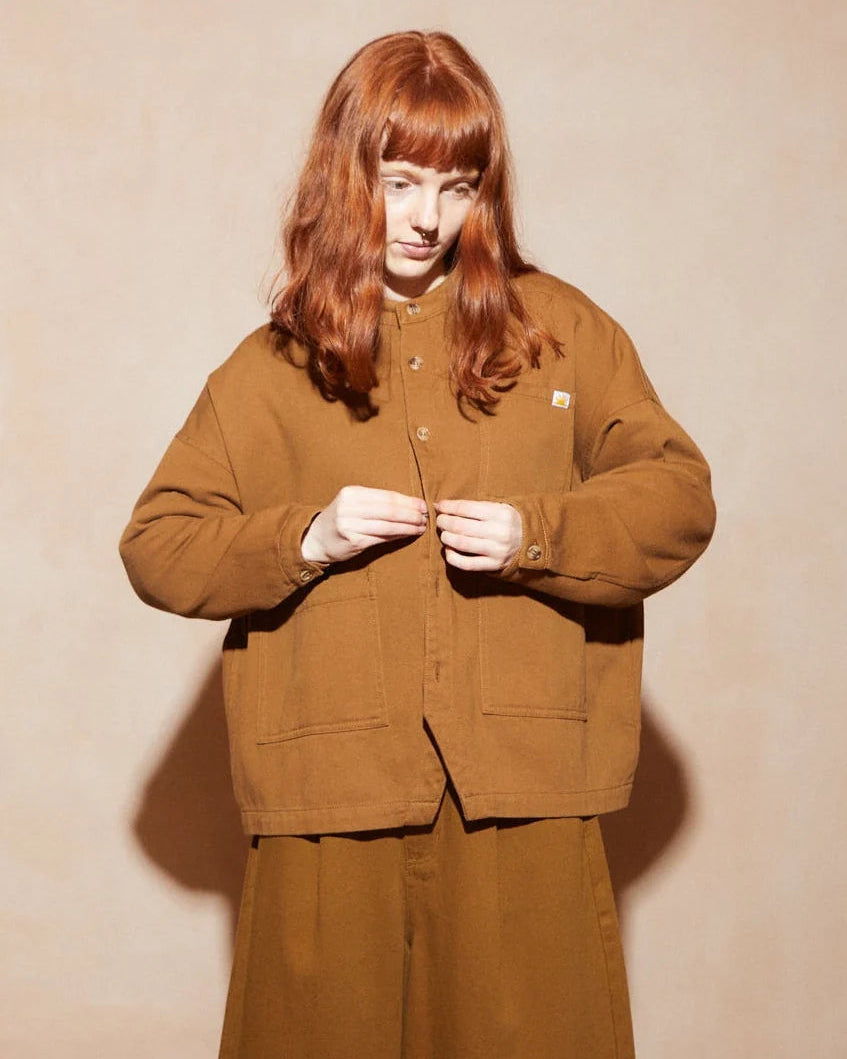 A model with long red hair wearing a brown corduroy jacket and matching wide-leg trousers, standing against a beige textured wall.