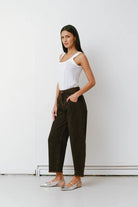 Woman wearing a white tank top and dark pants against a white wall