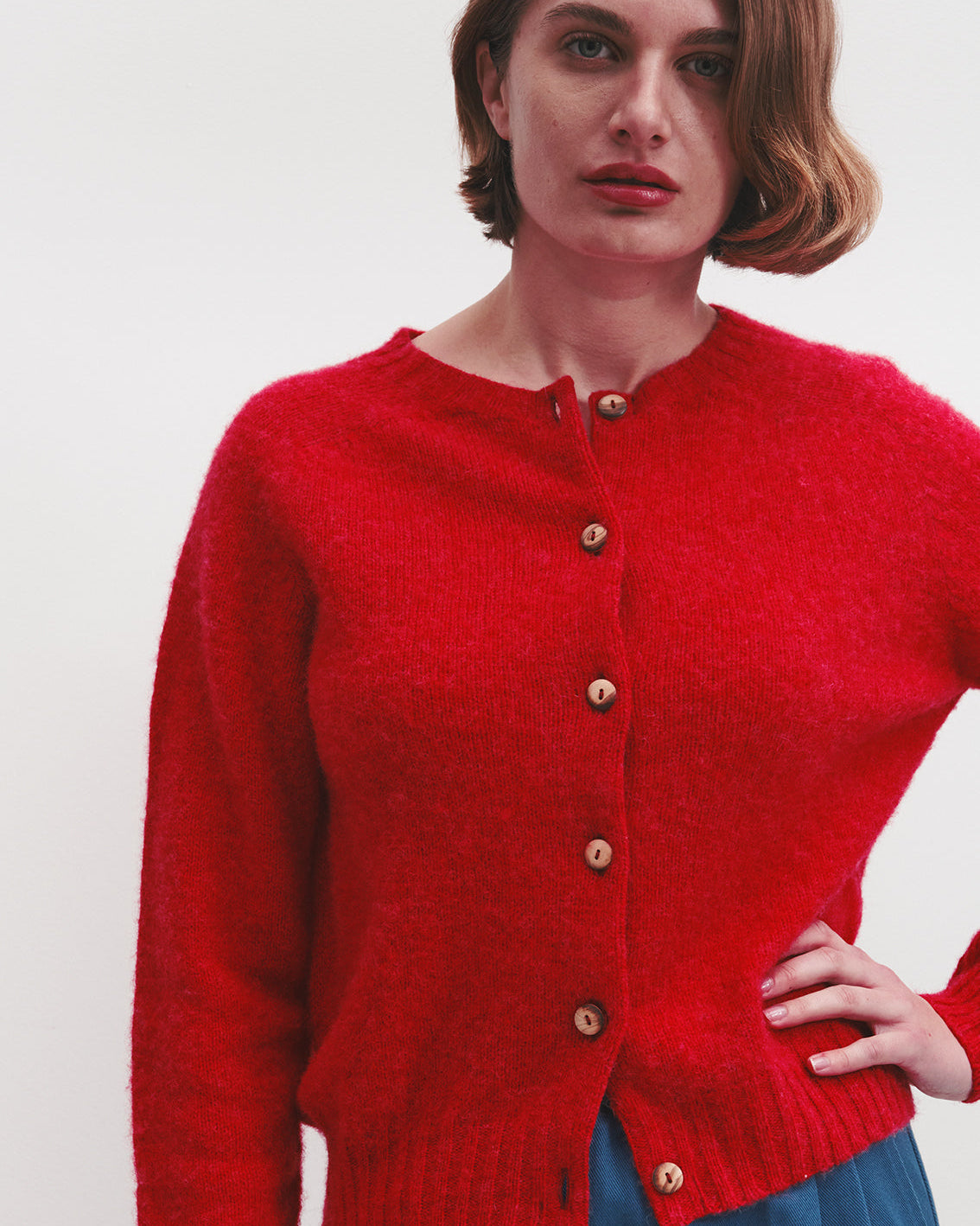 A woman wearing a bright red knit cardigan with brown buttons, paired with a blue skirt, photographed against a plain light background.