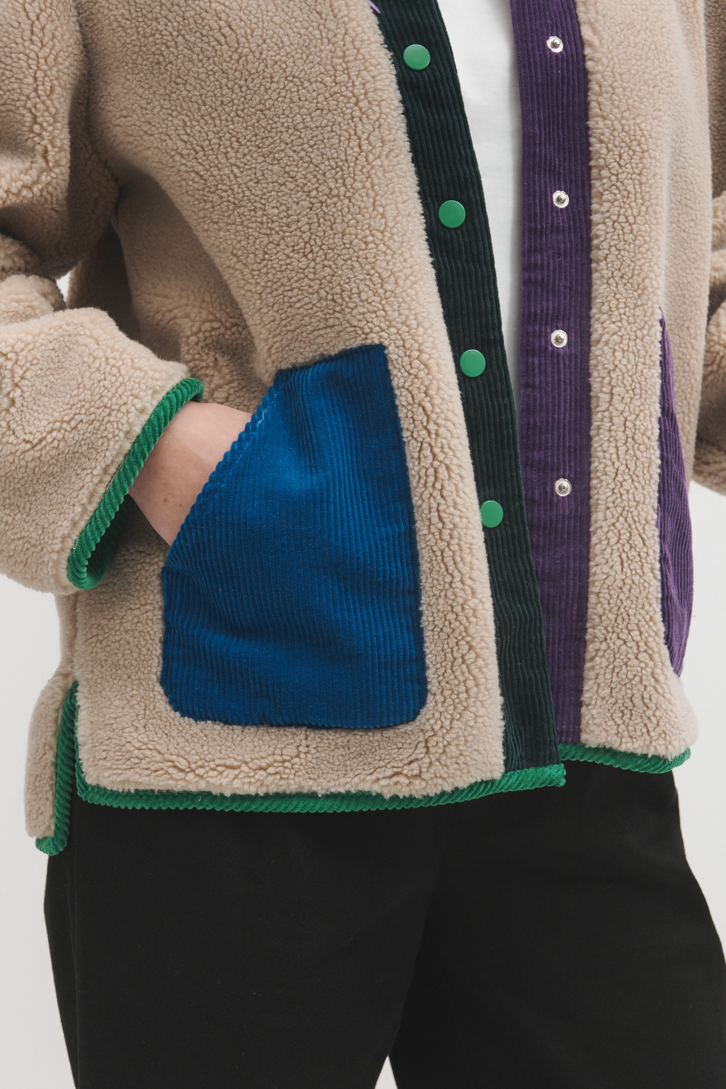 Close-up of a person wearing a beige sherpa cardigan with contrasting green trim, a blue corduroy front pocket, and purple corduroy side panels with green buttons.