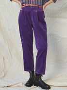 Person wearing purple corduroy pants with a checkered shirt and black boots on a textured beige background