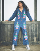 A woman stands outdoors against a weathered wooden fence wearing a blue long-sleeve jumpsuit with colorful printed motifs and white sneakers.