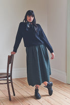 Woman wearing a navy sweater and green plaid skirt standing in a room with wooden floor and white walls.