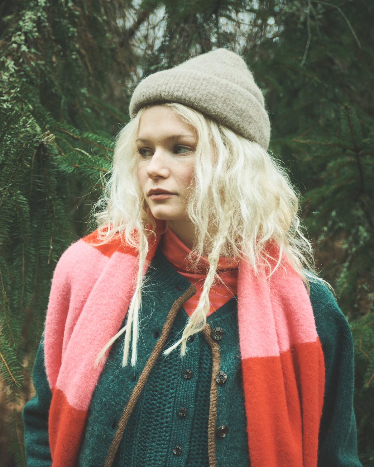 A woman standing outdoors in a forest setting, wearing a beige knit beanie, a dark green button-front cardigan, a pink and coral color-blocked knit sweater draped over her shoulders, and rust-red pants.