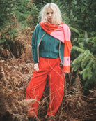 A model stands outdoors in a forest setting wearing a teal knit cardigan, bright red wide-leg pants, a pink and orange striped knit scarf, and a green top.