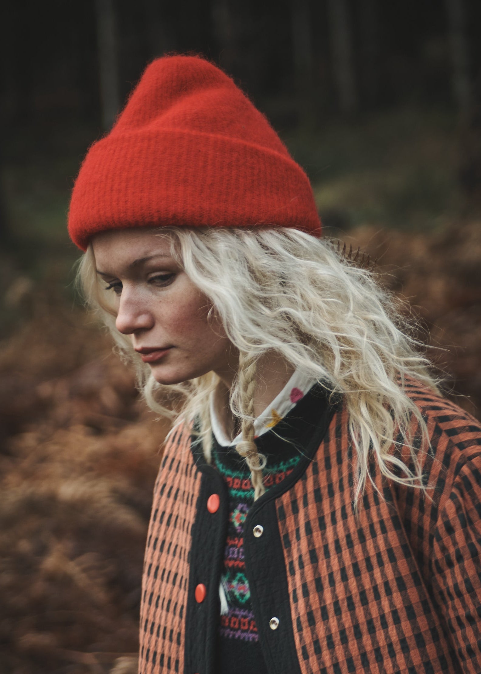 A woman wearing a bright red knit beanie and an orange-and-black checkered jacket, standing outdoors in a dark, wooded setting.
