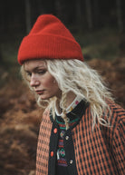A woman wearing a bright red knit beanie and an orange-and-black checkered jacket, standing outdoors in a dark, wooded setting.