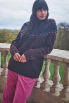 Woman wearing a dark sweater with colorful patterns and pink pants standing on a stone balcony.